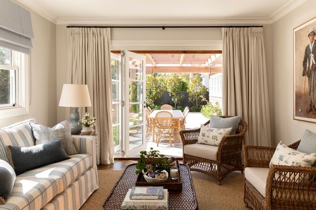 Bright, traditionally styled living room with a striped upholstered sofa, wicker armchairs, a central coffee table, neutral curtains, and framed artwork. French doors open onto a sunny outdoor dining terrace or patio, with a timber pergola visible beyond. The room features warm beige tones, layered textiles, and abundant natural light.