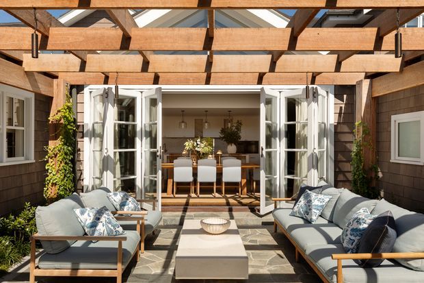 A bright outdoor living courtyard or covered terrace framed by a substantial timber pergola with exposed beams and translucent roof panels. Comfortable outdoor sofas and armchairs surround a low rectangular coffee table on patterned paving, while open French doors connect directly to an indoor dining area. The dark shingle-clad facade, white-trimmed glazing, climbing greenery, and warm timber structure create a refined indoor-outdoor entertaining space.
