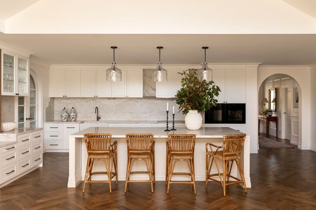 Bright, refined traditional kitchen with a large white island and four woven timber bar stools. The room features white Shaker-style cabinetry, a pale marble-look stone splashback and benchtops, dark herringbone timber flooring, arched openings and glazing, glass pendant lights, and classic decorative trims. An adjoining hallway and cabinetry are visible through the arched opening.