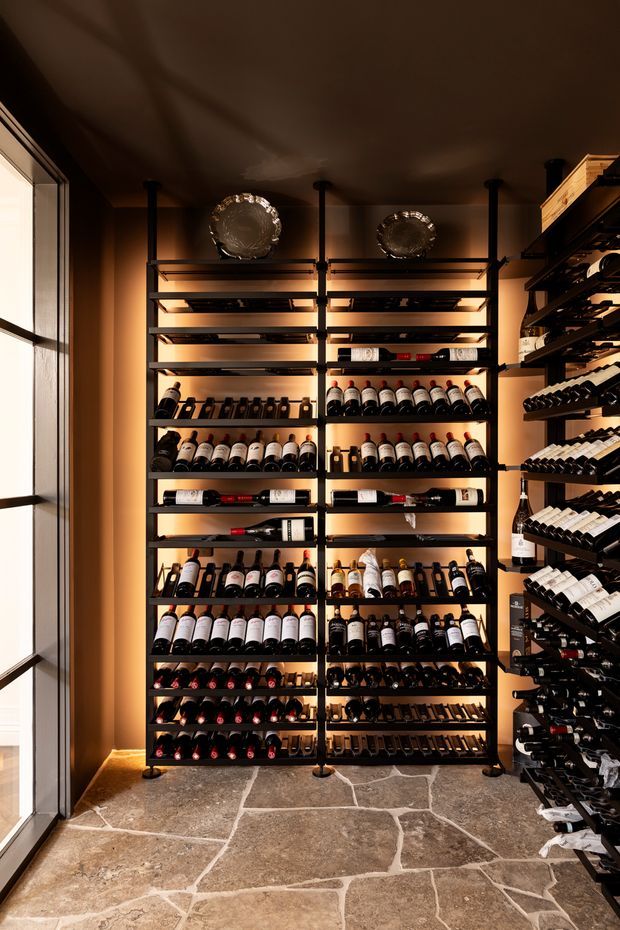 A warmly lit wine storage room with extensive black metal wine racks filled with bottles along the back and right walls. The room has a dark ceiling, warm tan walls, large window glazing on the left, decorative metallic platters on the upper shelving, and an irregular natural stone floor in earthy brown and grey tones.