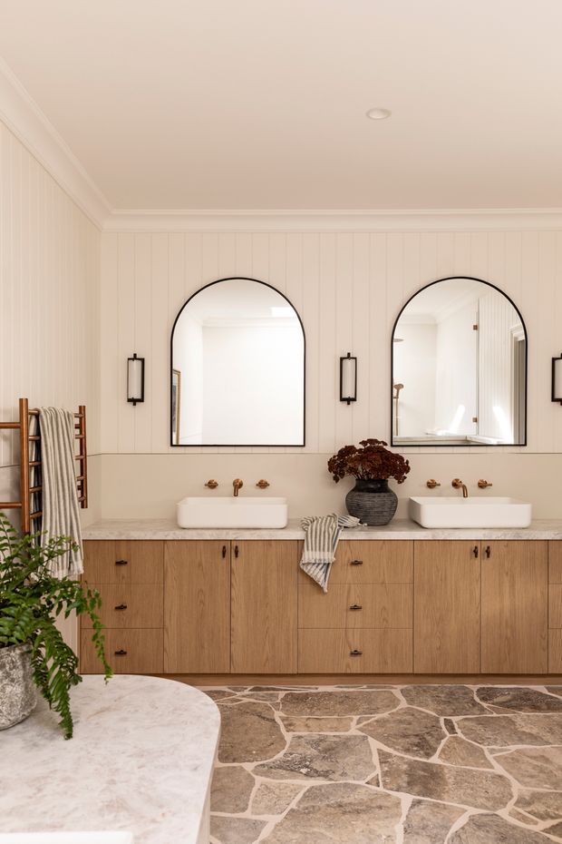 A warm, refined double-vanity bathroom with a long timber vanity, two white vessel basins, brass wall-mounted taps, arched black-framed mirrors, vertical wall panelling, and slim black wall sconces. The floor is finished in irregular natural stone slabs with light grout. A heated towel rail, towels, and potted greenery add functional and decorative detail.