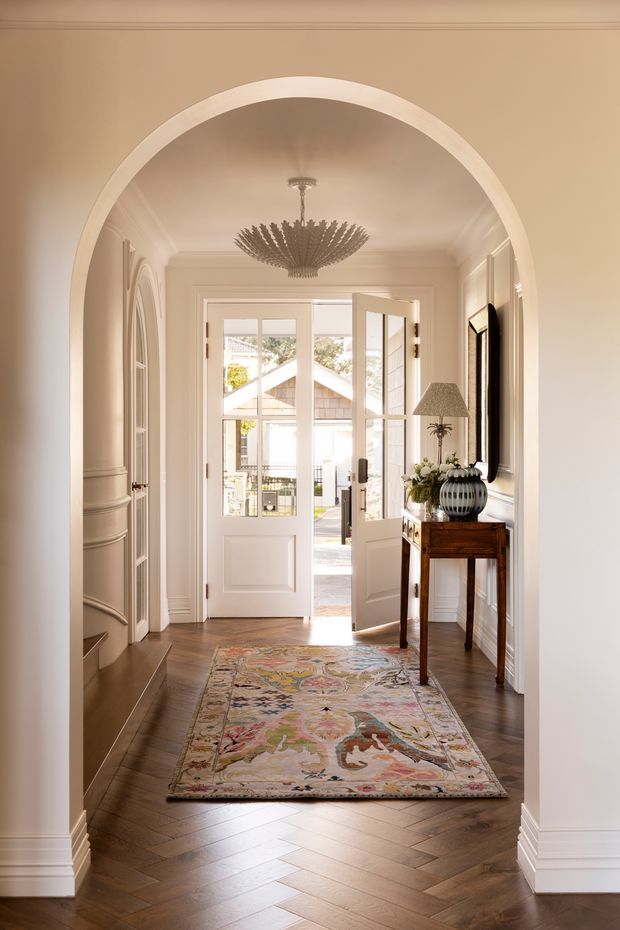 A refined residential entry hall viewed through a large rounded archway. The space features warm herringbone timber flooring, white painted panelled walls and trim, an ornate crystal chandelier, a patterned runner rug, a narrow dark wood console table with decorative objects, and a glazed front door opening to the exterior. Built-in arched wall detailing and classic mouldings create an elegant, traditional character.