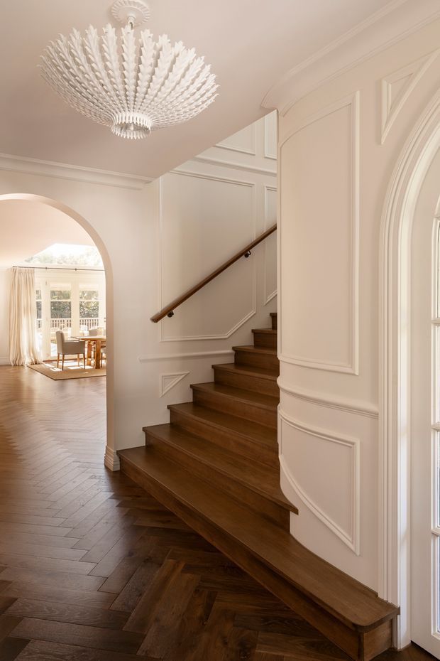 Elegant residential stairway and hallway with dark timber stairs and herringbone flooring, pale cream walls with extensive decorative panel moulding and curved arch details, a dark timber handrail, and a dramatic white feather-like pendant chandelier. An arched opening leads to a bright dining room with large windows and light curtains.