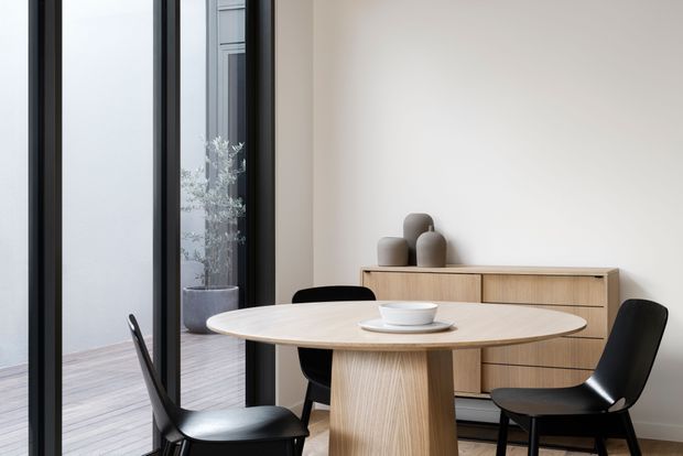 A refined contemporary dining area with a round light-wood dining table and sculptural pedestal base, surrounded by black dining chairs. A matching timber sideboard with rounded drawer fronts sits against a warm off-white wall, styled with neutral ceramic vessels. Full-height black-framed glazing opens to a narrow balcony or exterior space, bringing in soft natural light.
