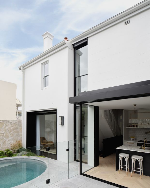 A contemporary two-storey white rendered home viewed from an outdoor courtyard and pool area. The facade combines crisp white walls, black-framed full-height glazing, a projecting dark beam, and a glass balustrade. Through the open glazing, a black-and-white kitchen with island seating and a staircase is visible. A small curved pool and pale stone paving occupy the foreground.