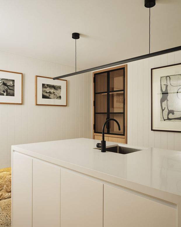 A refined contemporary kitchen centered on a large white island with integrated sink and matte-black gooseneck tap. The island has flat-panel white cabinetry and a smooth white benchtop. Vertical white wall panelling, framed monochrome artwork, a timber-framed black-grid window, and minimalist black pendant lights create a calm, warm modern interior.