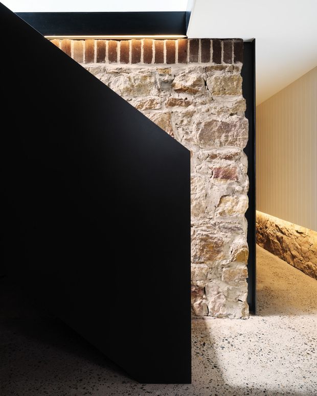Interior architectural detail featuring a bold matte-black geometric partition or stair-like volume in front of an exposed historic masonry wall. The wall combines rough pale stone with a red-brown brick course, while a terrazzo floor and warm concealed lighting continue into the adjacent corridor. The composition contrasts raw heritage materials with crisp contemporary black joinery and vertical wall panelling.