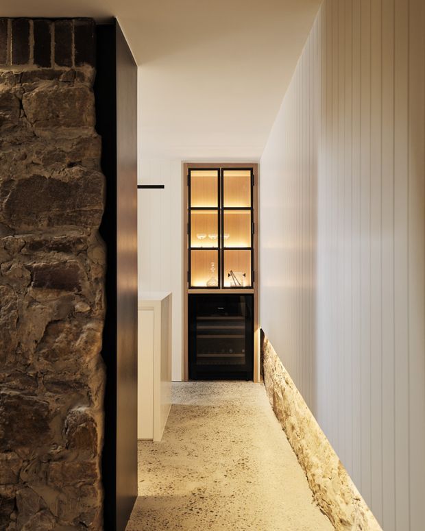 A narrow, warmly lit interior passage featuring a rough exposed stone wall on the left, vertical white wall panelling on the right, and a polished concrete or terrazzo-like floor. At the far end is a black-framed illuminated display cabinet with glassware and a built-in wine refrigerator below. The space has a refined rustic-contemporary character with strong contrast between natural stone, pale timber panelling, dark joinery, and warm lighting.