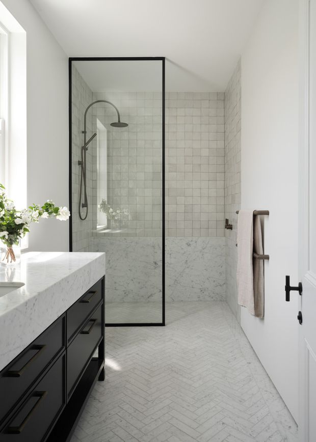 Bright, refined bathroom with a large walk-in shower enclosed by a slim black-framed glass screen. The shower walls feature pale square tiles above marble-look wall cladding, complemented by a white marble vanity top, black built-in cabinetry, black fixtures, and a light herringbone floor.