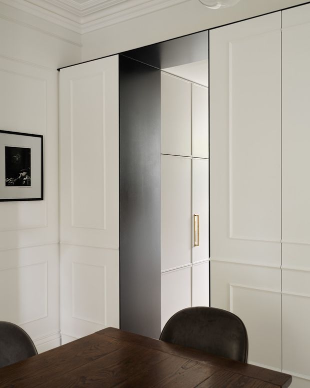 Elegant dining area with a dark timber dining table and upholstered chairs, surrounded by white decorative wall panelling and a large integrated built-in storage or wardrobe wall. A mirrored central recess and brass-toned vertical handle add contrast, while ornate ceiling cornices and framed artwork contribute a refined classic character.