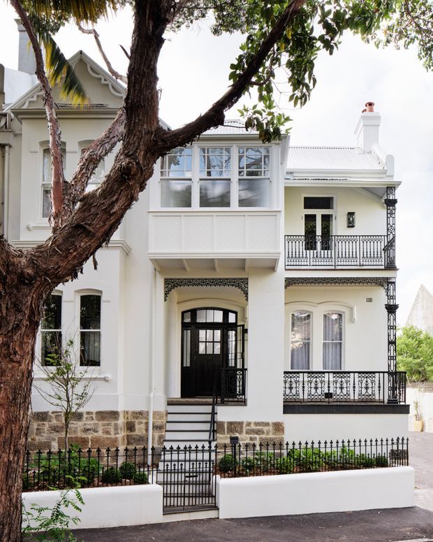 Front exterior view of a refined multi-storey heritage-style residence with a white rendered facade, ornate black wrought-iron balconies and lacework, tall sash-style windows, a projecting enclosed upper veranda, a pitched metal roof, and a small landscaped front garden behind a low white wall and iron fence. A large mature tree partially frames the facade.