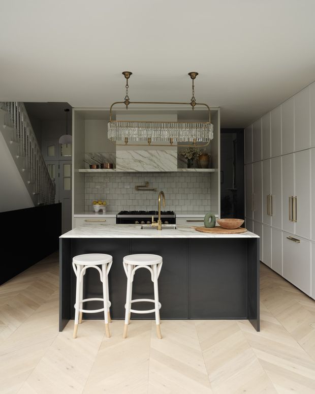 Refined kitchen interior with a large dark island, white marble-look benchtop, integrated sink and brass tapware, two white counter stools, pale herringbone timber flooring, white subway-tile splashback, extensive white cabinetry, and a decorative rectangular chandelier. A staircase and dark adjacent area are visible to the left.