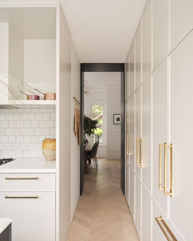 A narrow, refined kitchen passage leading into a dining area. White shaker-style cabinetry with brass handles lines the right wall, while the left side includes a white stone benchtop, white subway-tile splashback, open shelving, and a partially visible cooktop. A light timber herringbone floor extends toward a bright adjoining dining room with panelled walls, greenery, and a dark dining table. The overall scheme is bright, warm, and classic with contemporary detailing.