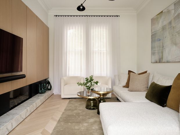 Refined contemporary living room with a large cream sectional sofa, sculptural coffee tables, pale herringbone timber flooring, a marble-lined feature media wall with warm timber cabinetry, sheer full-height curtains, and neutral artwork. The room is softly lit and layered with muted olive and tan accent cushions.