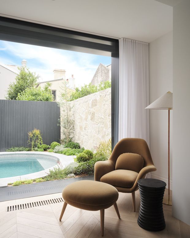 Bright, refined sitting area beside a large floor-to-ceiling window overlooking a small landscaped courtyard and swimming pool. The interior features a sculptural brown upholstered armchair and ottoman, a black ribbed side table, a slender floor lamp, white curtains, pale herringbone flooring, and dark window framing. An adjoining exterior courtyard with stone boundary walls, planting, and pool is visible through the glazing.