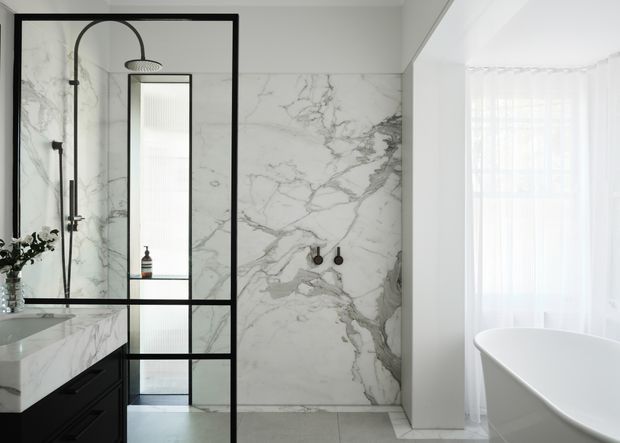 Elegant contemporary bathroom featuring a frameless-looking black metal-and-glass shower enclosure, dramatic white marble-look wall cladding with grey veining, a black vanity with a marble benchtop, and a freestanding white bathtub beside a bright sheer-curtained window.