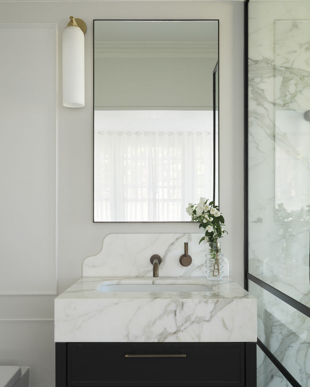 Elegant compact bathroom vanity with a white veined marble benchtop and integrated backsplash, black drawer cabinetry, brass wall-mounted fixtures, a frameless rectangular mirror, and a black-framed marble-look shower enclosure. Soft grey wall panelling and sheer reflected daylight create a refined, bright interior.