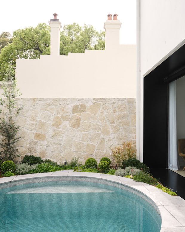 A compact landscaped courtyard with a curved tiled swimming pool in the foreground. The space is enclosed by a pale rendered wall topped with chimney-like forms and a lower natural stone-clad wall, with clipped shrubs and ornamental planting along the edges. A contemporary black-framed glazed opening is visible on the right, creating a strong contrast with the light masonry and pool.