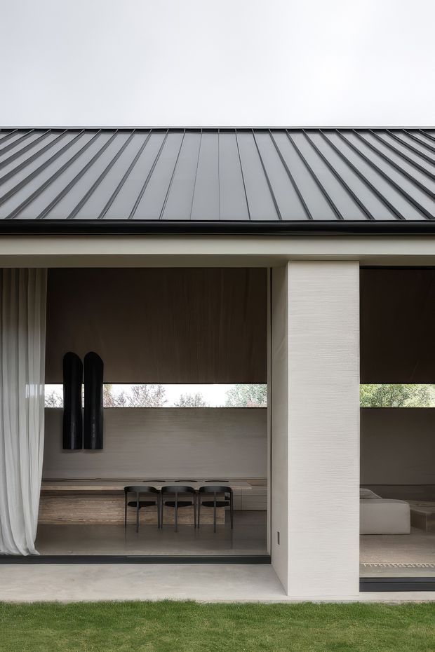 Exterior view of a refined contemporary residence with a dark standing-seam metal roof, broad recessed glazing, and a textured pale structural column. Through the glazing, a dining area and part of a living room are visible, featuring a long built-in bench, dark dining chairs, timber-toned walls, sheer curtains, and a restrained neutral palette. The house opens onto a narrow lawn.