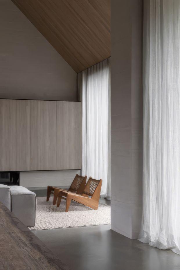 A refined, softly lit contemporary living room with a tall sloping timber-lined ceiling, full-height sheer curtains, pale vertical-grain built-in cabinetry, and smooth grey flooring. A pair of mid-century wooden lounge chairs sits on a light textured rug beside a partial light-coloured sofa. The space has generous vertical glazing, restrained materials, and a calm neutral palette.
