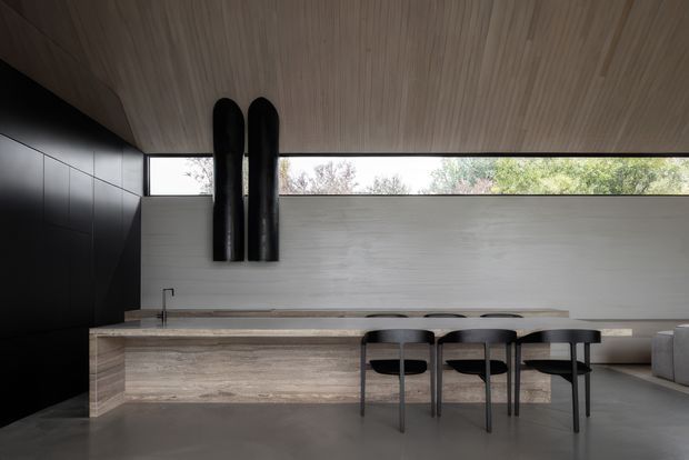 A minimalist open-plan kitchen and dining area with a long stone island and integrated seating. The room features dark matte cabinetry on the left, a pale textured stone island and backsplash wall, a timber-lined ceiling, a narrow clerestory window, and two tall black sculptural pendant lights. Four dark bar stools line the island, creating a refined, monochromatic interior with strong horizontal lines.