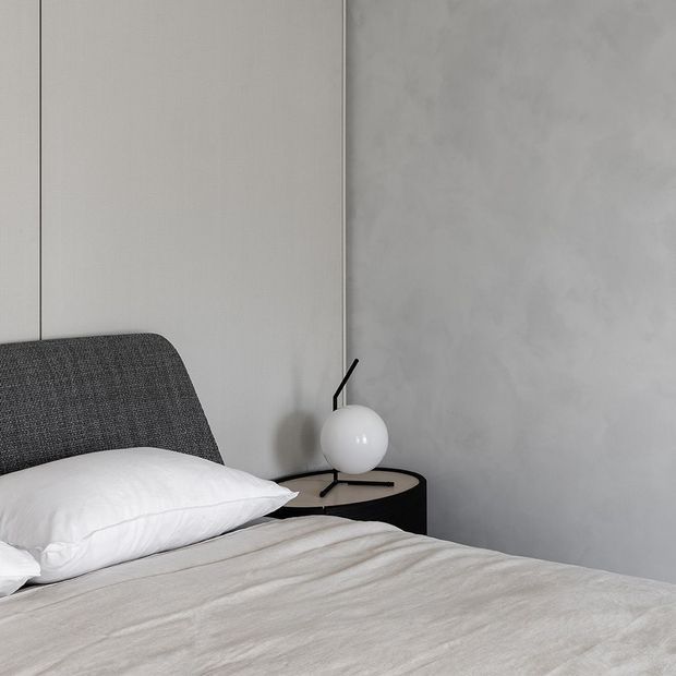 A minimalist bedroom vignette featuring a low upholstered dark grey bedhead, white bedding, a rounded black bedside table, and a white spherical lamp. The room has pale grey textured wall finishes and a restrained monochromatic palette.