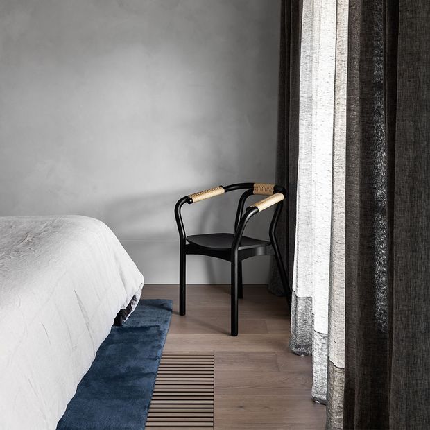 A minimalist bedroom corner with a neatly made bed, pale grey plastered walls, light timber flooring, a dark blue rug, and a black chair with woven cane arm details positioned beside layered sheer and dark curtains.