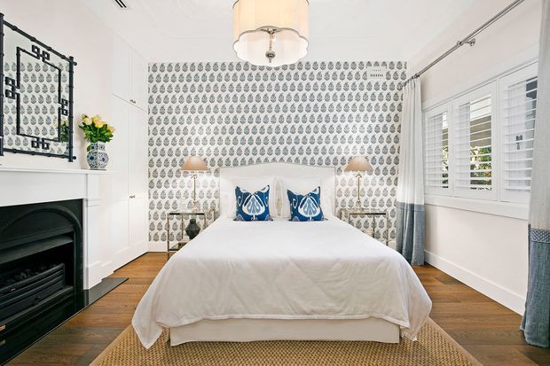 Bright, refined bedroom featuring a centered upholstered bed, patterned feature wallpaper behind the headboard, white built-in cabinetry, a dark fireplace surround, timber flooring, plantation shutters, and blue-and-white decorative accents. The room has a classic coastal feel with balanced symmetry and abundant natural light.