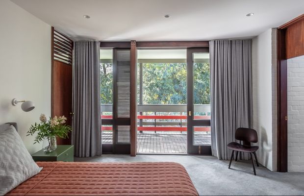 A refined bedroom with a low-profile bed in the foreground, upholstered in a dusty terracotta quilt. Full-height glazed doors open to a balcony, framed by dark timber detailing, louvred shutters, and full grey curtains. The room includes grey carpet, a small green bedside table, a wall-mounted reading light, and a dark timber chair. A white painted brick feature wall and warm timber built-in joinery add texture and contrast.