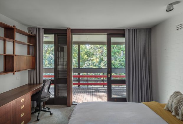 A contemporary bedroom with a built-in timber desk and open shelving along the left wall, a large bed in the foreground, and full-height glazed doors opening to a timber deck. Dark timber framing, grey curtains, a white painted brick wall, and leafy exterior views create a warm, calm atmosphere. A compact office chair and workspace are integrated into the bedroom.