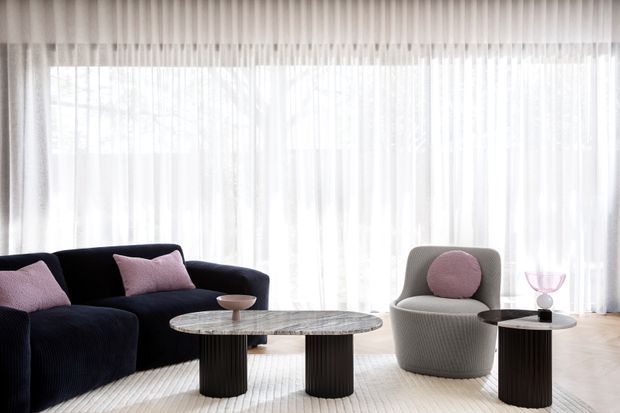 Bright, refined living room with a deep navy curved sofa, pink accent cushions, a rounded grey lounge chair, sculptural dark coffee tables, and full-height sheer curtains over expansive glazing. The room has a soft neutral palette and contemporary rounded furniture forms.