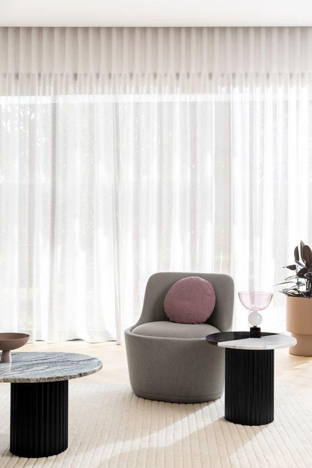 Bright, softly styled contemporary sitting area with full-height sheer curtains, a sculptural taupe upholstered lounge chair, round black fluted pedestal side tables, a pale stone-topped table, a blush circular cushion, and a potted plant. The room has light timber flooring and a large textured cream rug, creating an airy, refined atmosphere.