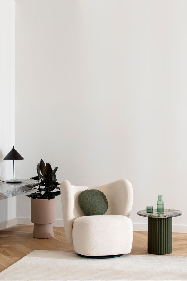 A refined minimalist living room vignette featuring a sculptural cream boucle armchair with a round green cushion, a dark green fluted side table with a marble-like top, a matching stone console, black pendant lamp, and potted plant. The room has white walls, warm herringbone timber flooring, and a pale textured rug.