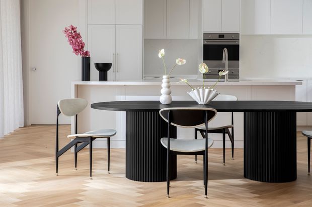 Bright contemporary open-plan kitchen and dining area with white full-height cabinetry, a pale stone island bench, and a sculptural black dining table with fluted pedestal bases. Light herringbone timber flooring, black-framed upholstered dining chairs, and restrained floral styling create a refined, minimalist interior.