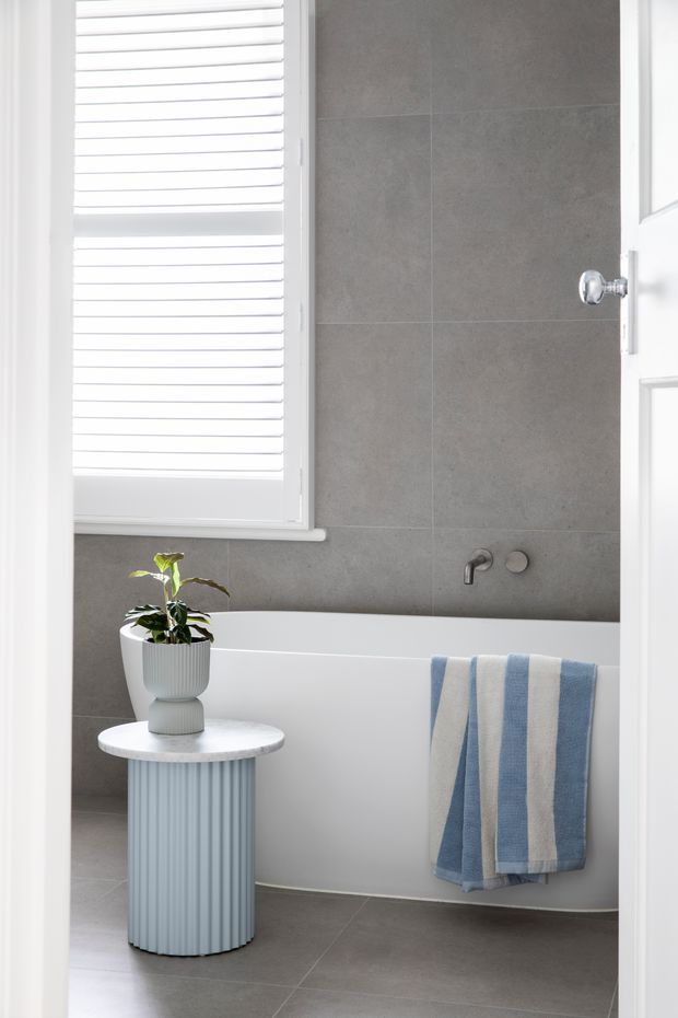 A bright, contemporary bathroom viewed through an open white door. The room features a freestanding white bathtub with wall-mounted chrome fixtures, large grey tiled walls and floor, a white shuttered window, and a small fluted side table holding a potted plant. A blue and white striped towel adds a subtle accent.