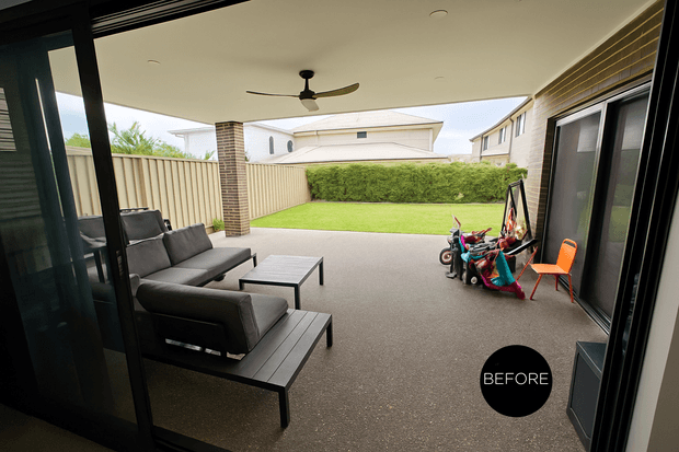 Covered outdoor patio viewed from inside through open glazed doors, with dark outdoor lounge furniture, a ceiling fan, tiled brick support column, and a broad paved entertaining area opening onto a fenced lawn and hedge. The image is marked “BEFORE,” indicating a pre-renovation or existing condition.