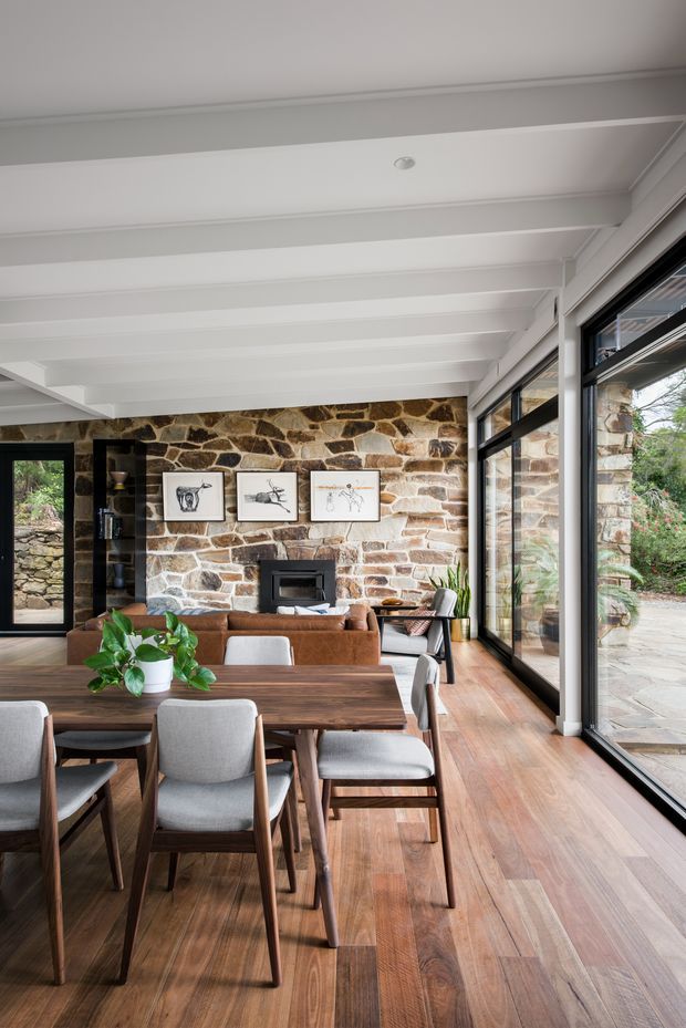 A bright open-plan dining and living area with a long dark timber dining table and upholstered chairs in the foreground. The room features warm timber flooring, a substantial natural stone fireplace wall with framed artwork, exposed white-painted ceiling beams, and expansive black-framed glazing opening toward a paved outdoor area. A brown sofa, lounge chair, built-in shelving, and indoor plants complete the warm, contemporary interior.