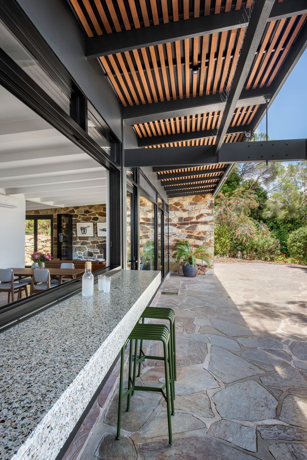 A covered outdoor entertaining area beside a glass-walled interior, featuring a long terrazzo-style stone bar benchtop with green bar stools, irregular natural stone paving, and a black steel pergola with warm timber slats overhead. Through the glazing, a dining area and stone feature wall are visible. Lush garden planting surrounds the sunlit patio.