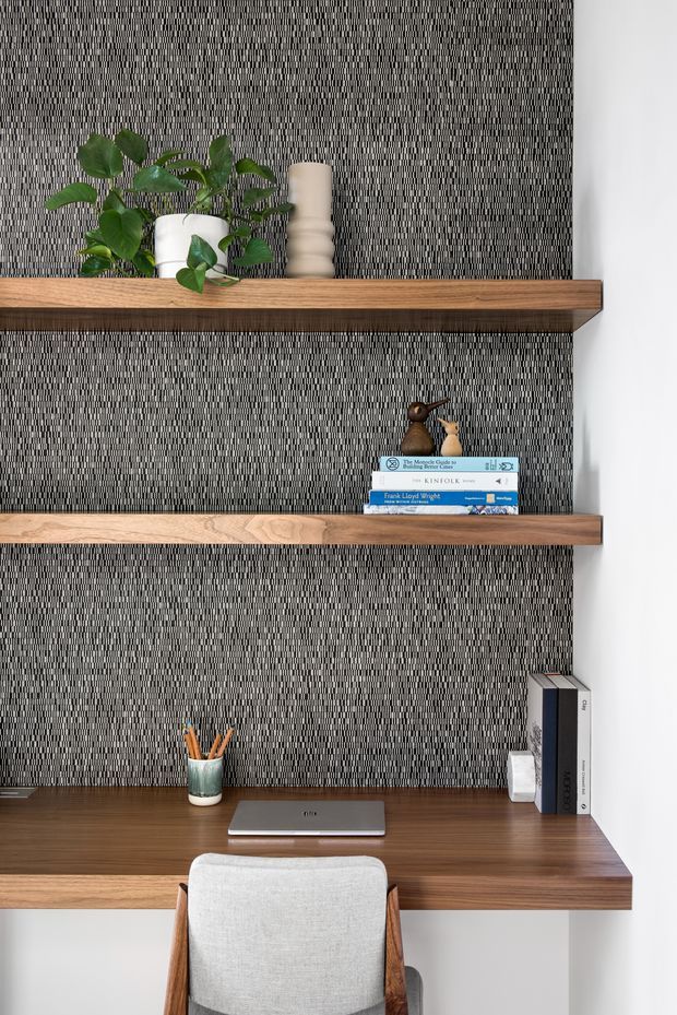 A compact built-in home workspace with a timber desk and two floating timber shelves set against a textured black-and-white vertical patterned wall. The desk includes a closed laptop, pencil cup, and books, while the shelves display a potted trailing plant, ceramic vessels, books, and small decorative objects. A light upholstered timber-framed chair sits at the desk. The overall look is refined, contemporary, and warmly monochromatic.