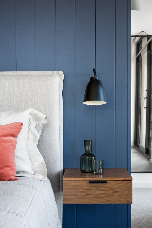 Close-up bedroom interior featuring a tall upholstered light-grey bedhead against a deep blue vertical-panelled wall. A black wall-mounted pendant hangs above a dark timber bedside table with black drawer pull and smoked glass vessels. White bedding and a coral-red cushion add contrast, while a mirrored wardrobe or glazed opening is visible at right.
