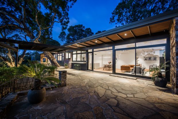 Twilight exterior view of a low-slung modern home opening onto a broad stone-paved courtyard or terrace. Extensive floor-to-ceiling glazing reveals a bright open-plan interior with dining and living areas, while a projecting roof and covered walkway create sheltered outdoor circulation. Dark metal framing, timber-lined soffits, exposed stone elements, mature trees and potted planting give the setting a refined, integrated indoor-outdoor character.