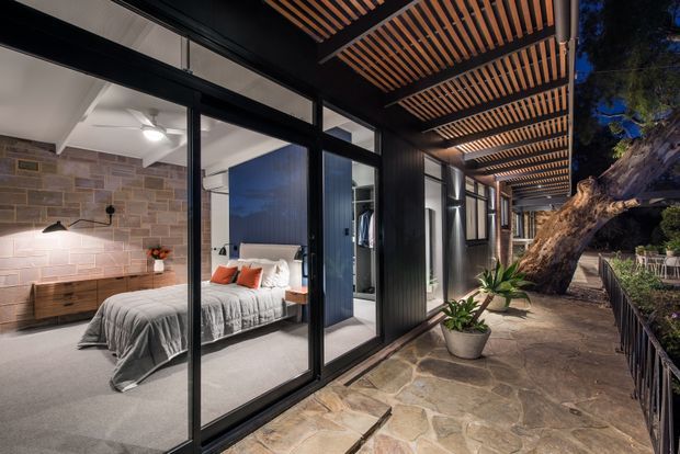A nighttime view of a modern bedroom opening onto a covered stone-paved outdoor walkway or terrace. Large black-framed glazed sliding doors connect the bedroom to the exterior, while a timber slatted pergola creates a strong linear ceiling overhead. The bedroom features a grey upholstered bed, white bedding with orange accent cushions, a timber bedside unit, pendant lighting, a ceiling fan, and a textured stone-look feature wall. Potted plants, mature trees, and outdoor seating are visible along the walkway.