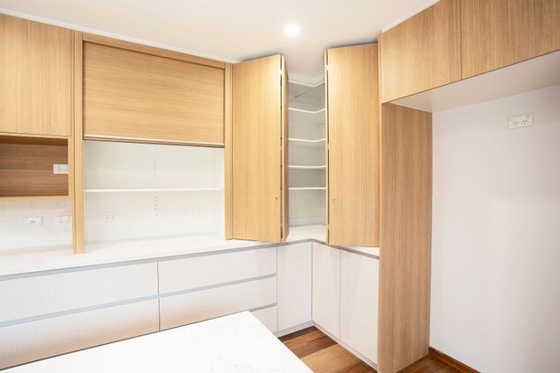 Bright built-in storage and work area with extensive light timber cabinetry, white drawers and benchtops, open shelving, and tall corner cupboards. A small portion of a white work surface is visible in the foreground, suggesting a compact home office or study nook. The room has warm timber flooring, white walls, recessed lighting, and a clean contemporary fit-out.