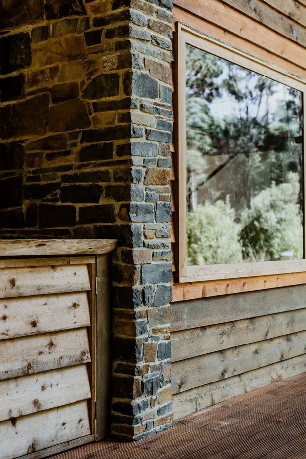 Close-up architectural detail of a rustic timber-clad exterior with a dark exposed brick corner, natural timber window frame, reflective glazing, and a timber deck. The weathered materials and surrounding vegetation visible through the window create a warm, cabin-like character.