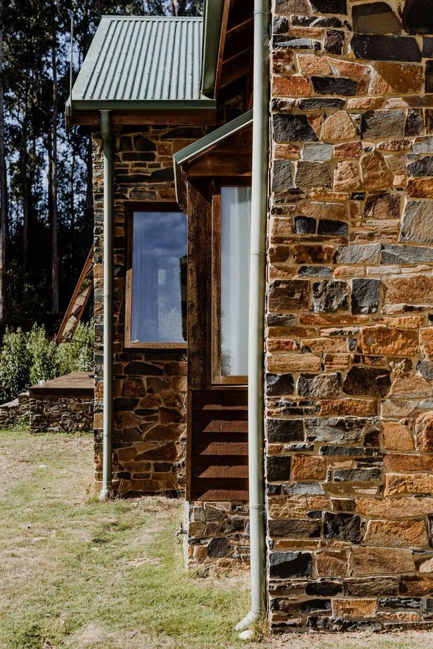 Close-up exterior view of a rustic stone building in a wooded setting. The facade uses irregular multicoloured natural stone masonry with dark timber window and door frames, corrugated metal roof sections, and exposed timber eaves. A narrow grassed area and adjacent stone structure are visible.