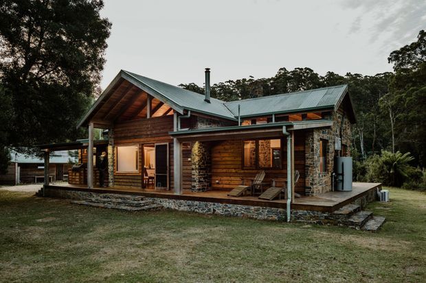 Exterior view of a rustic timber cabin set in a wooded landscape. The single-storey home has a pitched metal roof with multiple gables, exposed timber rafters and trusses, horizontal timber wall cladding, rough stone masonry piers and a raised covered veranda. Dark-framed windows, a chimney, outdoor seating and a rainwater tank are visible.