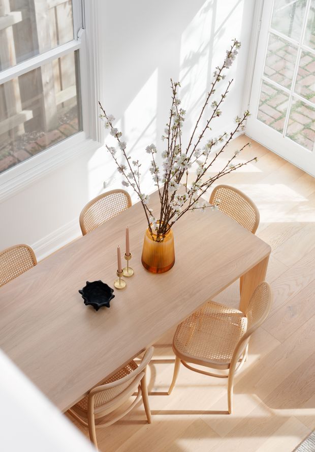 Bright, airy dining room viewed from above, featuring a light timber dining table surrounded by natural cane-back chairs. Large white-framed windows and glazed doors fill the room with sunlight, while a simple floral branch arrangement in an amber vase forms the centrepiece. Light timber flooring and white walls create a warm, refined contemporary setting.