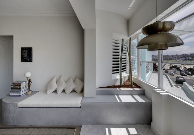 Bright contemporary interior corner with a built-in upholstered window seat and white cushions, adjacent to a large corner of glazing overlooking an urban waterfront or marina setting. A sculptural layered pendant light hangs above the space, while a narrow planter with tall architectural plants sits beside white louvred shutters. The palette is restrained in white, light grey and concrete tones, with strong sunlight and shadows creating an airy, refined atmosphere.