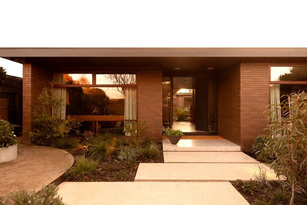 Front exterior of a low, single-storey brick residence with a broad flat roof overhang, deep recessed entry, timber-framed glazing, and pale concrete stepping paths. Established garden planting frames the entry and connects to a curved paved walkway. The warm-toned brickwork and horizontal composition create a restrained mid-century character.