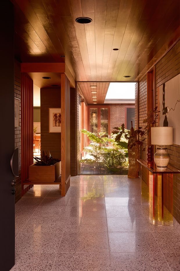 A warm, richly detailed residential hallway or entry passage opening onto an interior courtyard. The space features a glossy timber-lined ceiling, reddish-brown brick walls, polished terrazzo flooring, timber-framed glazing and doors, recessed ceiling lights, and built-in timber display shelving with decorative ceramics. Lush planting fills the bright courtyard beyond, with a skylight or roof opening bringing in daylight.
